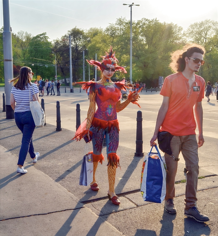 15 - A person showcasing intricate, colorful full-body paint resembling a phoenix or exotic bird, complete with a matching feathered headpiece and accessories while walking outdoors